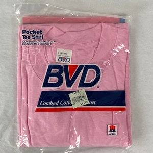 Könnte beinhalten: Ein pinkfarbenes T-Shirt mit Tasche, BVD-Logo und dem Schriftzug "Combed Cotton" in Blau und Rot. Das Shirt ist in Plastik verpackt und hat ein Größenetikett mit der Aufschrift "L (42-44)".