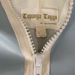K&ouml;nnte beinhalten: Nahaufnahme eines wei&szlig;en Rei&szlig;verschlusses mit einem wei&szlig;en Etikett, auf dem "Topanga Toggs of California" steht. Das Etikett enth&auml;lt auch den Text "Style Size 8 RN 26710". Der Rei&szlig;verschluss-Schieber tr&auml;gt das YKK-Logo.