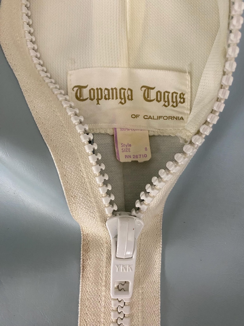 K&ouml;nnte beinhalten: Nahaufnahme eines wei&szlig;en Rei&szlig;verschlusses mit einem wei&szlig;en Etikett, auf dem "Topanga Toggs of California" steht. Das Etikett enth&auml;lt auch den Text "Style Size 8 RN 26710". Der Rei&szlig;verschluss-Schieber tr&auml;gt das YKK-Logo.