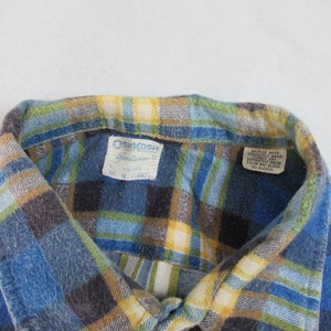 以下が含まれることがあります： 青、黄色、緑の格子柄のフランネルシャツで、OshKosh B'Goshのラベルが付いています。ラベルには「OshKosh B'Gosh」と「Sportwear」の文字が書かれており、サイズタグには「L」、「100% コットン」、「米国製」と記載されています。