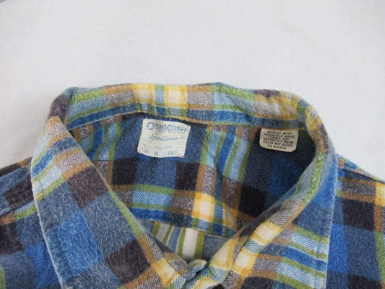 以下が含まれることがあります： 青、黄色、緑の格子柄のフランネルシャツで、OshKosh B'Goshのラベルが付いています。ラベルには「OshKosh B'Gosh」と「Sportwear」の文字が書かれており、サイズタグには「L」、「100% コットン」、「米国製」と記載されています。