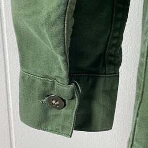 May include: Close-up of a green jacket sleeve. The sleeve features a buttoned cuff with a dark button. The fabric has a textured appearance, and the stitching is visible along the seams. The jacket appears to be made of a durable material.