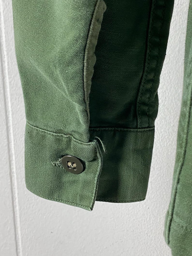 May include: Close-up of a green jacket sleeve. The sleeve features a buttoned cuff with a dark button. The fabric has a textured appearance, and the stitching is visible along the seams. The jacket appears to be made of a durable material.