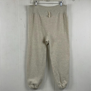 Może przedstawiać: Jasnoszare spodnie dresowe ze sznurkiem w pasie i elastycznymi mankietami. Spodnie wiszą na wieszaku na tle białej, panelowej ściany. Materiał wydaje się być miękką, wygodną tkaniną.