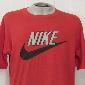 Puede incluir: Camiseta roja de manga corta con un logotipo de Nike en blanco y negro. El logotipo es un swoosh con la palabra "NIKE" en letras blancas.