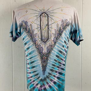 Puede incluir: Una camiseta tie-dye con un dise&ntilde;o de cristal en la espalda. La camiseta es de color azul claro con un patr&oacute;n tie-dye blanco y morado. El dise&ntilde;o de cristal est&aacute; en el centro de la espalda y presenta un cristal grande con cristales m&aacute;s peque&ntilde;os que lo rodean.