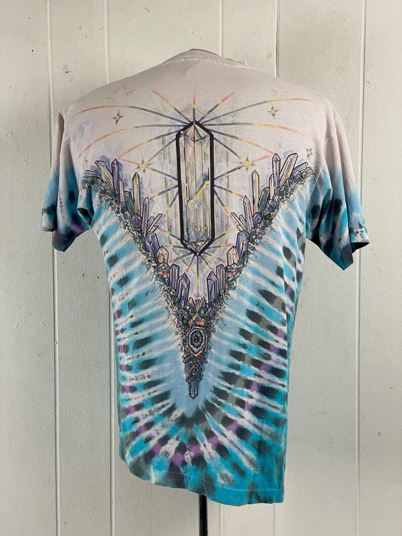Puede incluir: Una camiseta tie-dye con un dise&ntilde;o de cristal en la espalda. La camiseta es de color azul claro con un patr&oacute;n tie-dye blanco y morado. El dise&ntilde;o de cristal est&aacute; en el centro de la espalda y presenta un cristal grande con cristales m&aacute;s peque&ntilde;os que lo rodean.