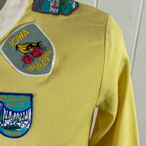 May include: Yellow long-sleeved shirt with various state and location patches. Patches include "Iowa, Tall Corn State", "Niagara Falls, Canada", "Vermont", and "Stratford, Connecticut".
