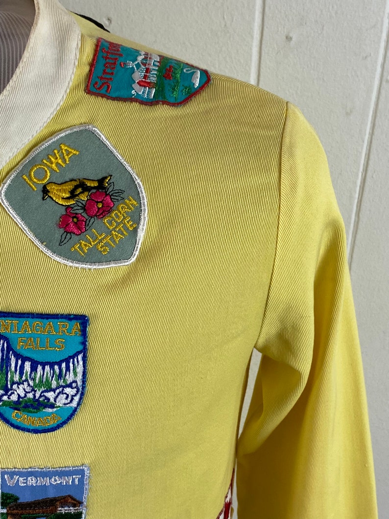 May include: Yellow long-sleeved shirt with various state and location patches. Patches include "Iowa, Tall Corn State", "Niagara Falls, Canada", "Vermont", and "Stratford, Connecticut".