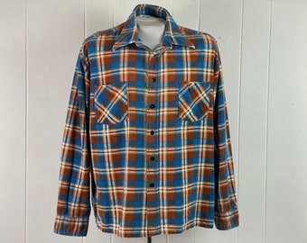 Può includere: Una camicia di flanella a maniche lunghe con un motivo a quadri blu, arancio e bianco. La camicia ha un colletto abbottonato e due tasche sul petto.