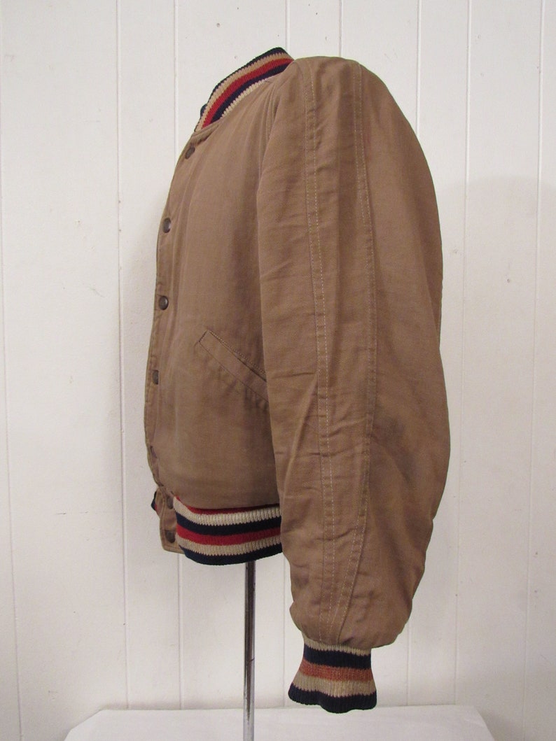 May include: A brown bomber jacket with a ribbed collar and cuffs. The cuffs have red, white, and blue stripes.