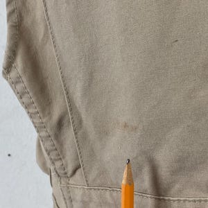 May include: Close-up of a tan-colored garment with visible stitching and a pencil. The fabric appears to be a sturdy material, possibly canvas or a similar durable textile. The pencil is orange with a black tip.