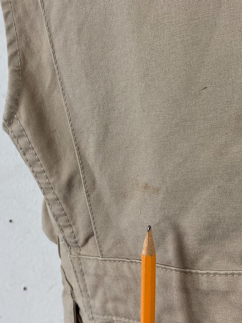 May include: Close-up of a tan-colored garment with visible stitching and a pencil. The fabric appears to be a sturdy material, possibly canvas or a similar durable textile. The pencil is orange with a black tip.