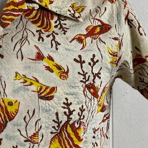 May include: A white short-sleeved shirt with a colorful fish and coral print. The shirt has a button-down collar and a pocket on the left side.
