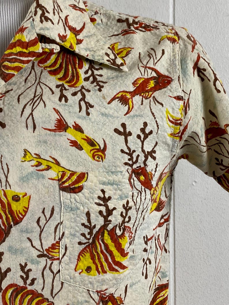 May include: A white short-sleeved shirt with a colorful fish and coral print. The shirt has a button-down collar and a pocket on the left side.