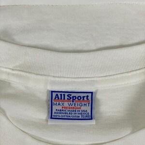 May include: Close-up of a white t-shirt with a blue and white tag. The tag reads "All Sport MAX WEIGHT PRESHRUNK FABRIC MADE IN USA ASSEMBLED IN MEXICO 100% COTTON/COTON XL(48)". The shirt is made of cotton.