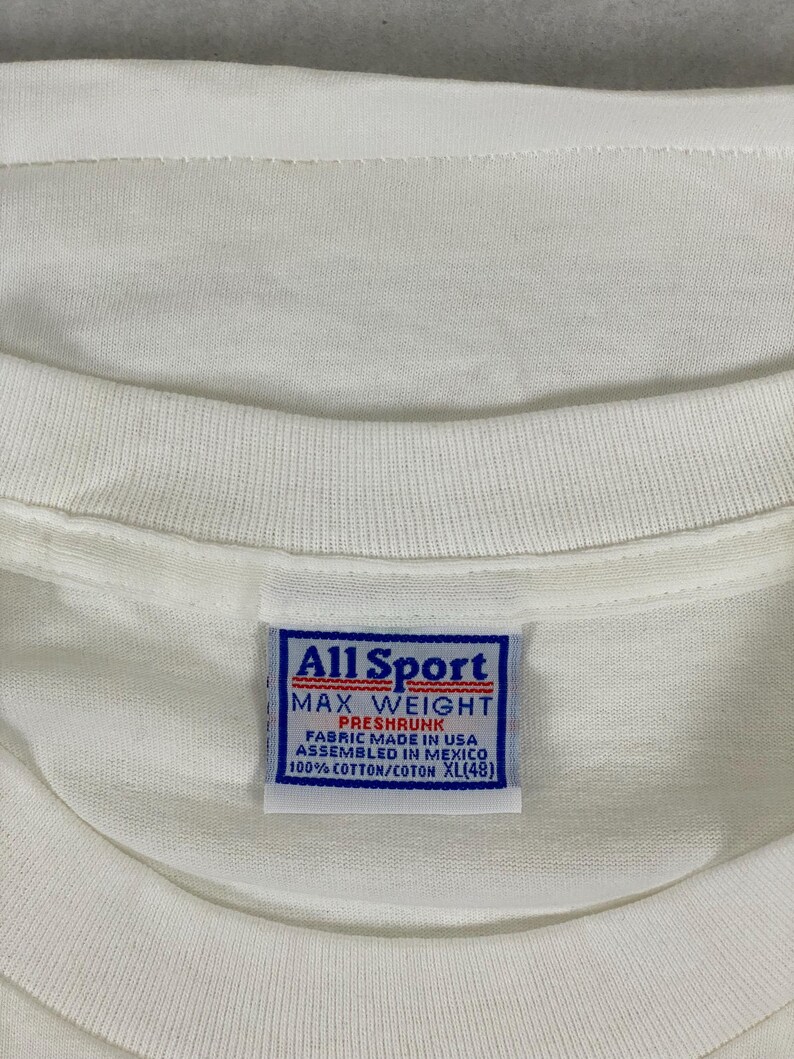May include: Close-up of a white t-shirt with a blue and white tag. The tag reads "All Sport MAX WEIGHT PRESHRUNK FABRIC MADE IN USA ASSEMBLED IN MEXICO 100% COTTON/COTON XL(48)". The shirt is made of cotton.