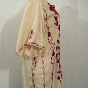 May include: A white tunic with red embroidered floral designs. The tunic has a loose fit and a round neckline.