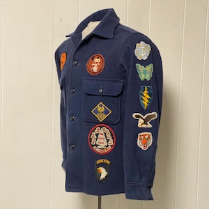 May include: A navy blue jacket adorned with various embroidered patches. Patches feature designs such as Mickey Mouse, a butterfly, a lightning bolt, and a tiger. The jacket has two chest pockets and button fastenings.