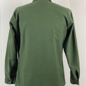 May include: Olive green long-sleeve shirt with a button-down front and a collared neckline. The shirt has buttoned cuffs and a classic, utilitarian design. The back of the shirt is visible, showcasing the tailored fit and durable fabric.