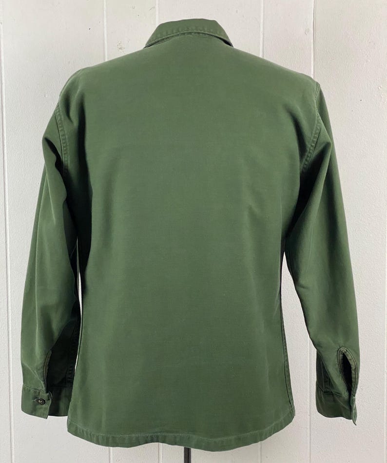 May include: Olive green long-sleeve shirt with a button-down front and a collared neckline. The shirt has buttoned cuffs and a classic, utilitarian design. The back of the shirt is visible, showcasing the tailored fit and durable fabric.