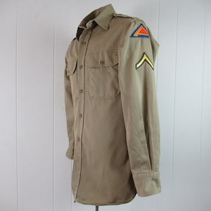 Può includere: Una camicia a maniche lunghe color beige con una toppa sulla manica sinistra. La toppa è un triangolo rosso, giallo e blu con un chevron giallo sulla manica destra.