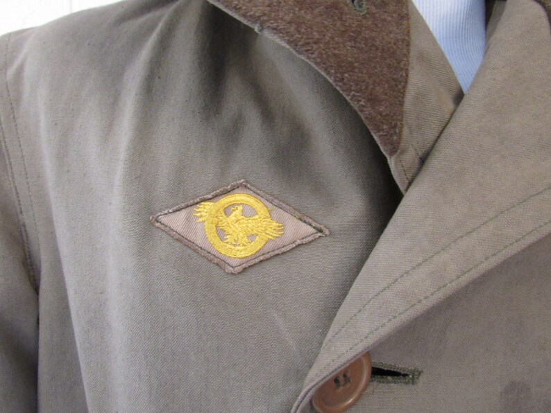 May include: A brown jacket with a brown corduroy collar and a gold eagle patch on the left side of the chest.