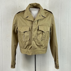 May include: A tan military-style jacket featuring a pointed collar, long sleeves with buttoned cuffs, and two large chest pockets with button-down flaps. The jacket has shoulder epaulets and a straight hemline, constructed from a durable cotton fabric.
