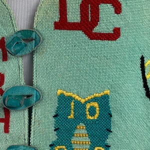 May include: A teal blue knitted vest with colorful embroidered letters and a cartoon cat design. The vest has turquoise buttons.