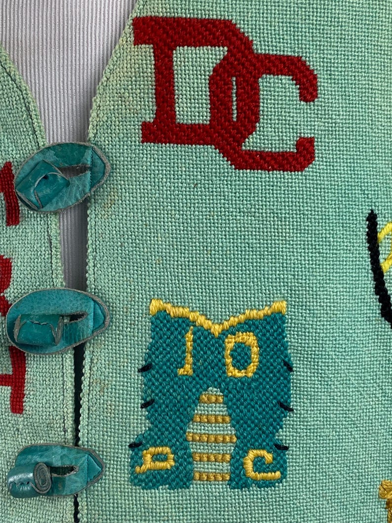 May include: A teal blue knitted vest with colorful embroidered letters and a cartoon cat design. The vest has turquoise buttons.