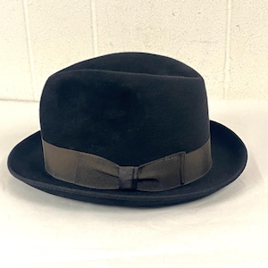 May include: A black fedora hat with a dark brown ribbon band and bow. The hat has a rounded crown and a wide brim. The hat is sitting on a white surface.