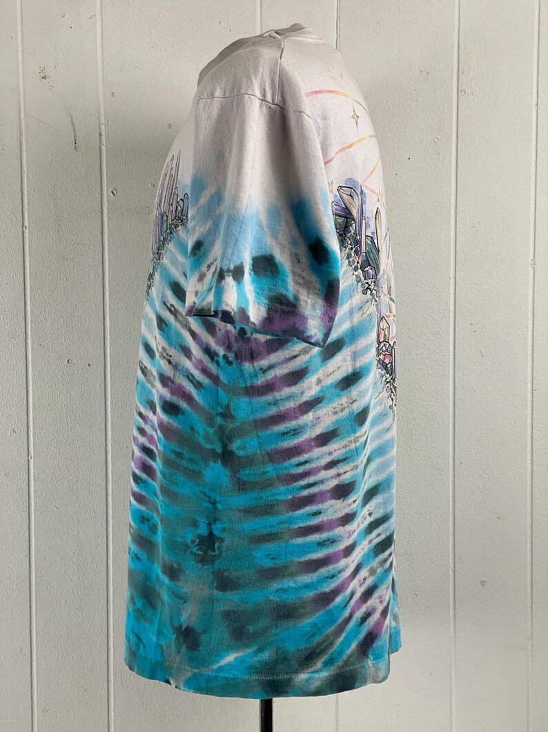 Puede incluir: Una camiseta blanca tie-dye con un patr&oacute;n de remolino azul, morado y verde. La camiseta presenta una impresi&oacute;n gr&aacute;fica de una ciudad con un arco&iacute;ris y estrellas.