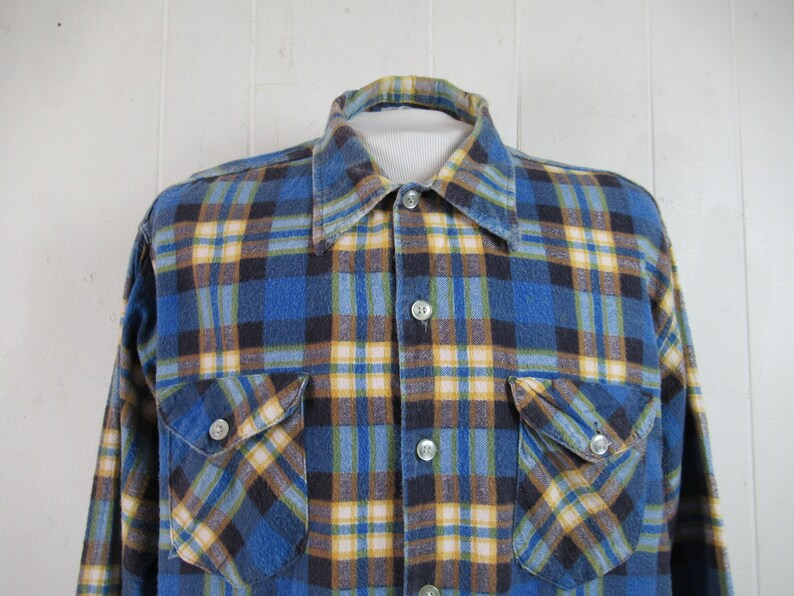 以下が含まれることがあります： 長袖で胸ポケットが2つ付いた、青、黄、黒の格子柄のフランネルシャツです。