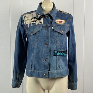 Puede incluir: Una chaqueta vaquera azul con parches, incluyendo un parche de piel sintética en el hombro izquierdo, un parche de "Wonder Bread" en el pecho derecho y un parche de "The Doors" en el pecho izquierdo.