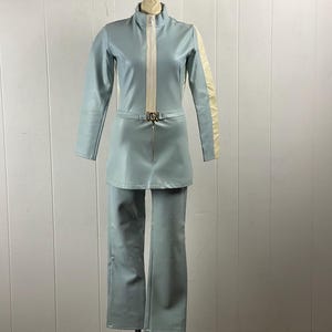K&ouml;nnte beinhalten: Ein hellblauer Jumpsuit mit einem wei&szlig;en Streifen an jeder Seite der &Auml;rmel. Der Jumpsuit hat einen Rei&szlig;verschluss vorne und einen G&uuml;rtel mit einer Metallschnalle.