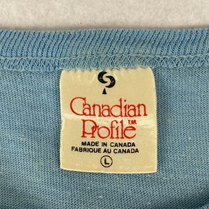 K&ouml;nnte beinhalten: Ein wei&szlig;es Etikett mit dem Text "Canadian Profile" in roten Buchstaben. Das Etikett tr&auml;gt au&szlig;erdem die Aufschrift "Made in Canada Fabrique au Canada" und ein Gr&ouml;&szlig;enetikett "L".