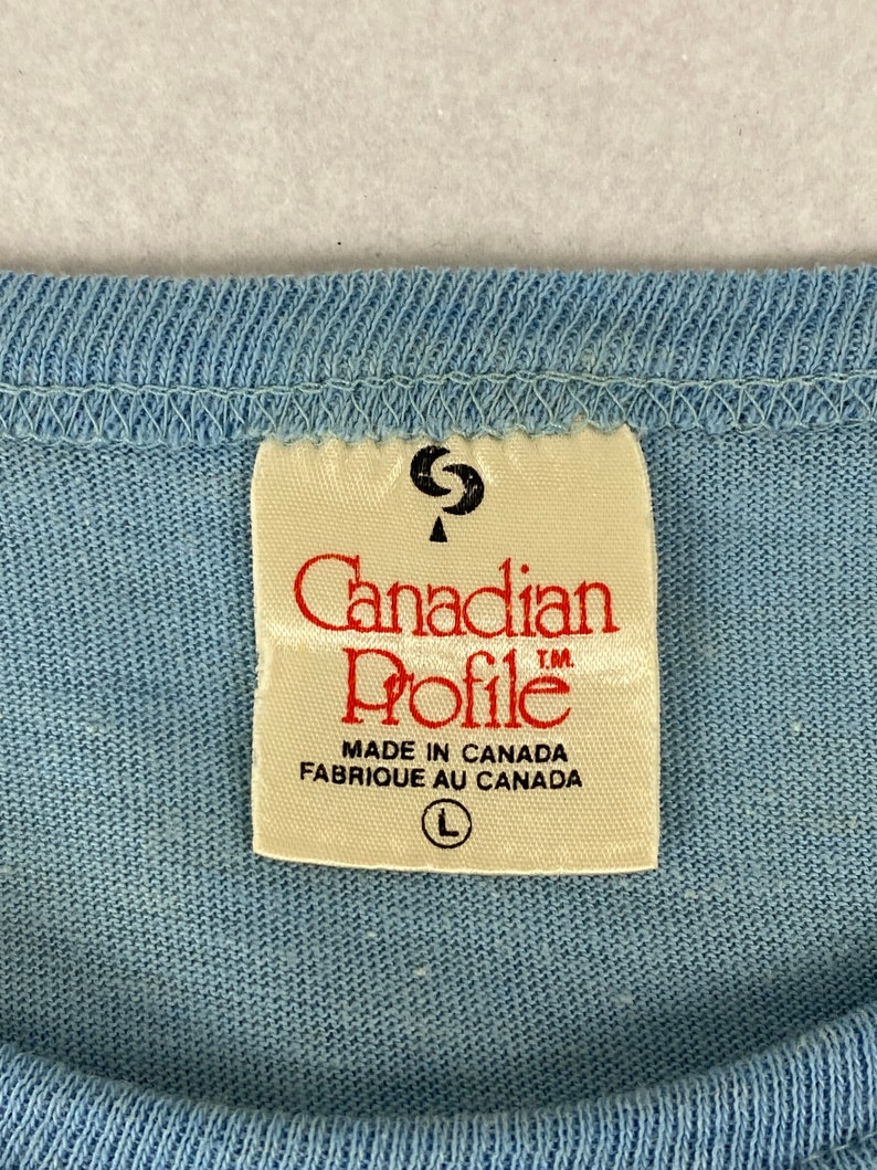 K&ouml;nnte beinhalten: Ein wei&szlig;es Etikett mit dem Text "Canadian Profile" in roten Buchstaben. Das Etikett tr&auml;gt au&szlig;erdem die Aufschrift "Made in Canada Fabrique au Canada" und ein Gr&ouml;&szlig;enetikett "L".
