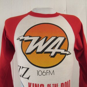 Op de afbeelding: Een wit en rood langarm baseballshirt met een grote gele en oranje cirkelvormige afbeelding met de tekst "W4 106FM" en de tekst "King of the Road" onder de cirkel.
