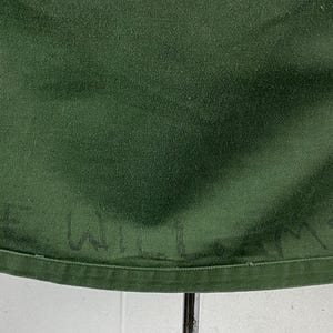 May include: Close-up of a dark green fabric with the handwritten name "E. WILLIAMS" visible near the bottom edge. The fabric has a slightly textured appearance and is likely part of a garment.