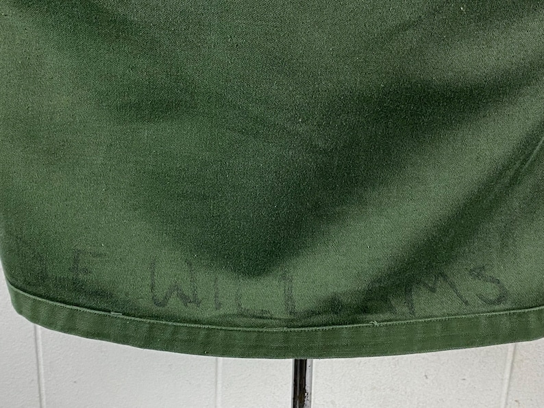 May include: Close-up of a dark green fabric with the handwritten name "E. WILLIAMS" visible near the bottom edge. The fabric has a slightly textured appearance and is likely part of a garment.