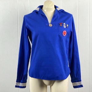 May include: A royal blue long-sleeved shirt with a sailor collar and a small pocket. The collar and cuffs have white stripes. The shirt has a decorative wheel emblem and several pins on the chest.