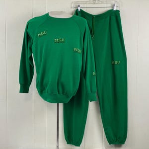 Puede incluir: Un conjunto verde de sudadera y pantalón de chándal a juego. La sudadera y el pantalón tienen las letras "MSU" bordadas en dorado. La sudadera es de manga larga con cuello redondo. El pantalón de chándal tiene cintura con cordón.