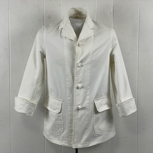 Puede incluir: Chaqueta blanca con cuello, tres botones y dos bolsillos delanteros. Las mangas son de tres cuartos con puños doblados. La chaqueta es de tela clara y cuelga de una percha sobre un fondo blanco.