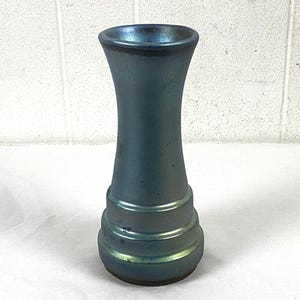 Könnte beinhalten: Eine blau schillernde Glasvase mit ausgestellter Oberseite und einer Reihe horizontaler Rippen nahe der Basis. Die Vase hat eine glatte, glänzende Oberfläche und einen subtilen Farbwechsel von Blau nach Grün.