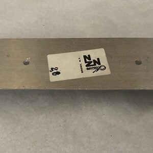 May include: A silver-coloured metal bracket, rectangular with a U-shaped bend. It features two small holes and a sticker with "NZ Masson Bronx, N.Y." and the number "28".