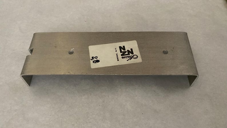 May include: A silver-coloured metal bracket, rectangular with a U-shaped bend. It features two small holes and a sticker with "NZ Masson Bronx, N.Y." and the number "28".
