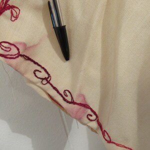 May include: Close-up of a cream-colored fabric with a red embroidered design. The fabric has a subtle texture and the embroidery is intricate.