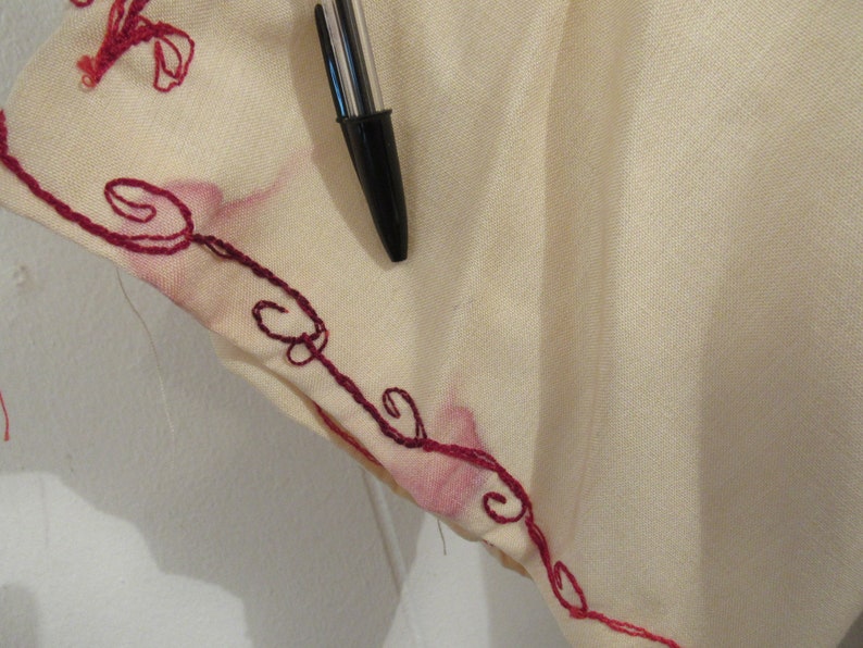 May include: Close-up of a cream-colored fabric with a red embroidered design. The fabric has a subtle texture and the embroidery is intricate.