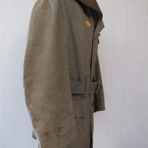 May include: A brown and tan military style jacket with a brown collar and a belt. The jacket has a patch on the left side of the chest with a yellow emblem.