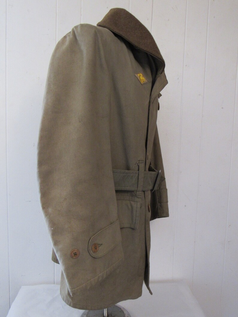 May include: A brown and tan military style jacket with a brown collar and a belt. The jacket has a patch on the left side of the chest with a yellow emblem.
